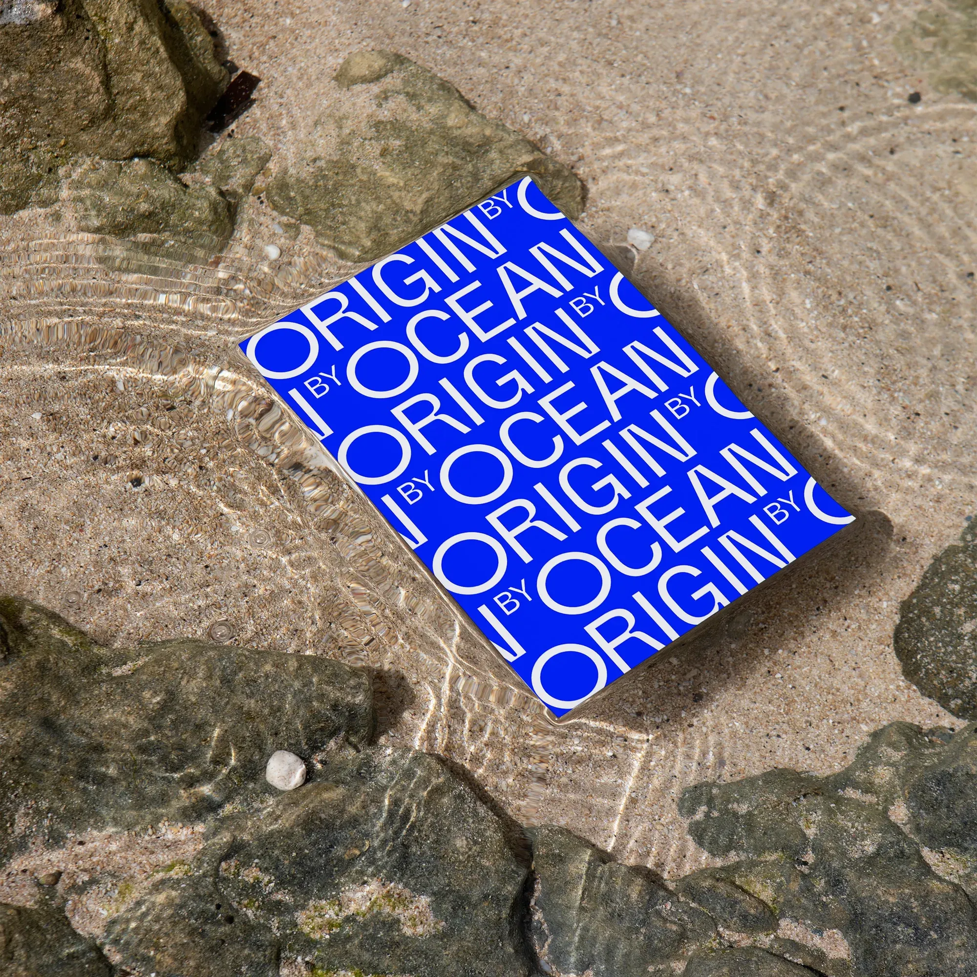 Origin by Ocean brand book with a repeated blue logotype, lying in shallow seawater on rocks.