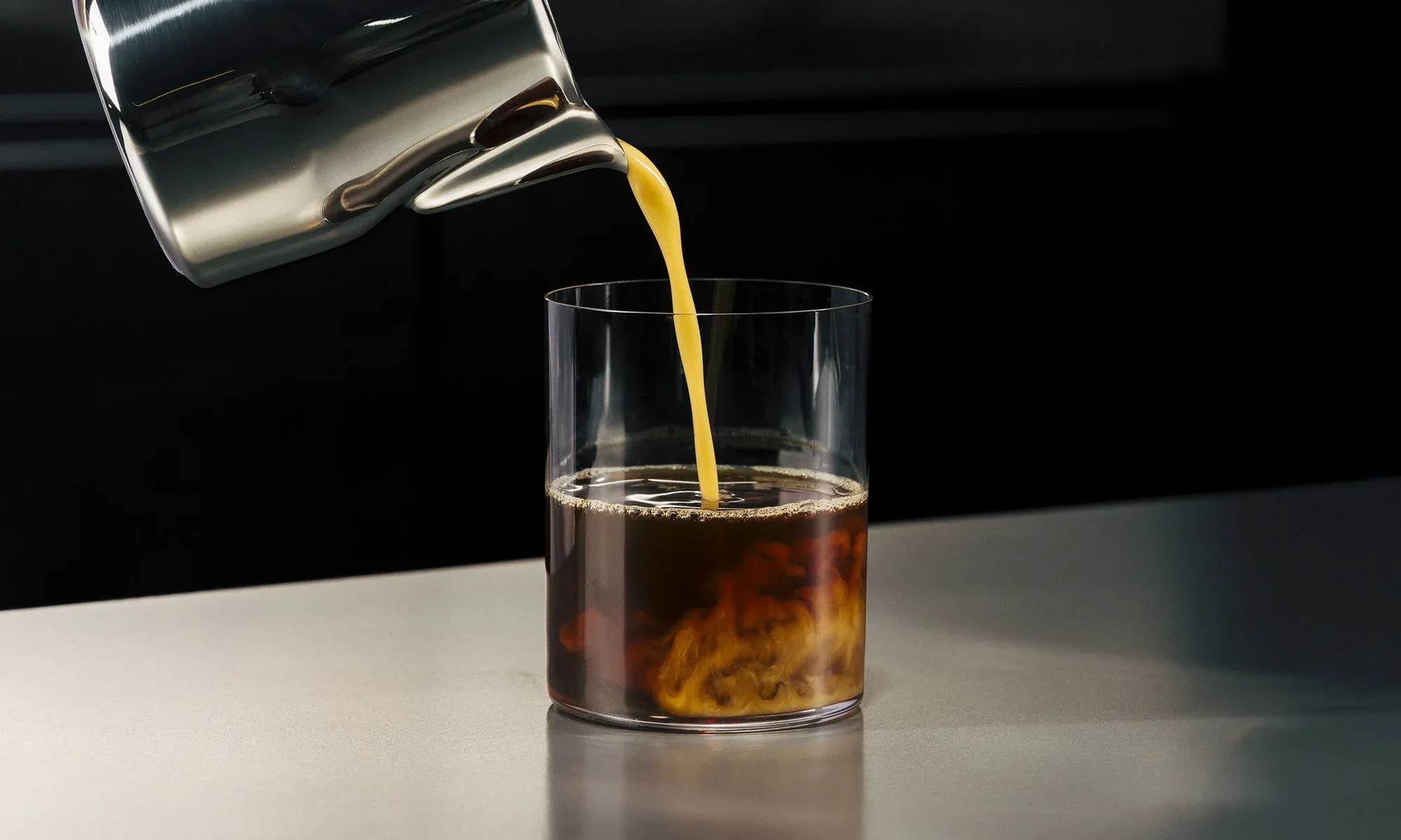 Cream poured from a steel jug into a glass of iced coffee.